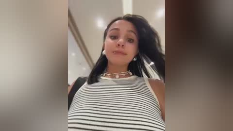 Video of a young woman with medium skin tone, straight black hair, and a sleeveless striped top, standing indoors with blurred background.