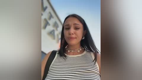 Video of a young woman with long black hair, wearing a striped tank top, standing in front of a white building with a blue sky background.
