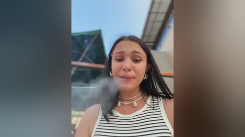 Video of a young woman with long black hair, wearing a striped tank top, smoking a cigarette, standing outdoors with a blurred background.