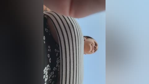 Video of a young woman with short dark hair, wearing a black and white striped shirt, looking upward against a clear blue sky.