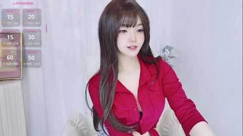 Video of a young Asian woman with long, straight black hair, fair skin, and wearing a red blouse, sitting in a white room with a radiator and a temperature display on the left.