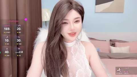A video of an Asian woman with long, dark hair in a white lace top, smiling. She stands in a modern bedroom with pink bedding, blue walls, and a digital camera overlay.