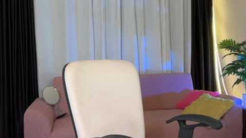 A video of a modern living room with a white office chair, a pink sofa, black curtains, and a yellow pillow; a green potted plant in the background.