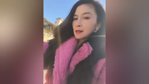 A video of a young Asian woman with long, dark hair, wearing a pink and black fur coat, smiling outdoors under a clear blue sky with a rustic stone building in the background.