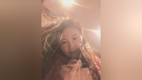 Video of a young Asian woman with long, disheveled hair, wearing a pink jacket, captured in a dimly lit room with a warm, soft glow.