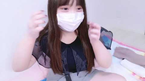 Video of an East Asian woman with long brown hair, wearing a black lace blouse, white mask, and gray skirt, sitting cross-legged on a colorful blanket. She appears to be adjusting her mask.