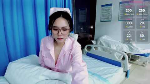 Media: Video of an Asian woman in a pink nurse's uniform and cap, sitting on a hospital bed, wearing glasses. Hospital room with blue curtains, another bed, and a digital screen displaying vital signs.