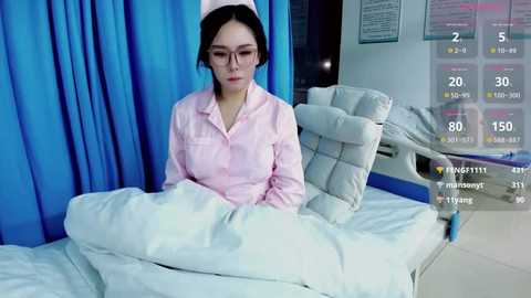 Video of a young Asian woman with glasses, wearing a pink hospital gown, sitting on a white bed in a sterile hospital room with blue curtains and digital medical monitoring screens.