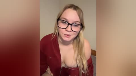 Video of a fair-skinned, blonde woman with glasses, wearing a red robe and beige lace lingerie, leaning forward with a slight smile.