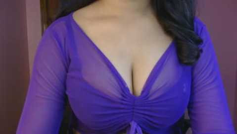 Media: Video of a woman with medium skin tone wearing a sheer, purple, long-sleeved top that reveals her cleavage, highlighting her large breasts. Her dark, wavy hair falls over her shoulders. The background is a muted, pinkish wall.