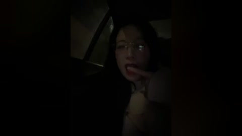 A dimly lit video of a woman with long black hair and glasses, wearing a black coat, with a hand to her mouth, in a dark, shadowy car interior.