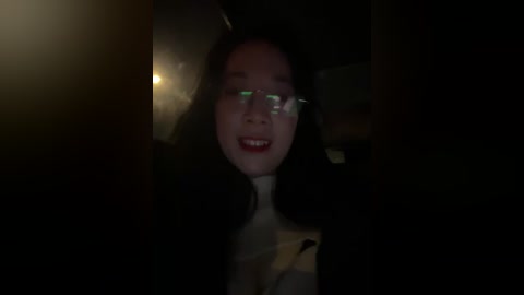 Video of a smiling Asian woman with long black hair, wearing glasses and a white shirt, in a dimly lit, possibly urban setting.