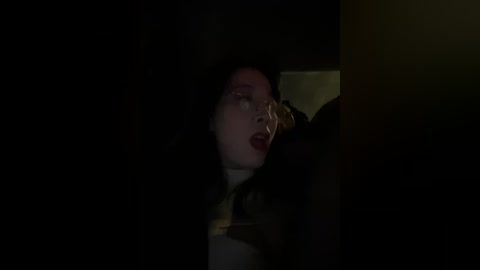 A dimly lit video of a woman with pale skin and dark hair, wearing red lipstick, lying on a bed, partially obscured by shadows.