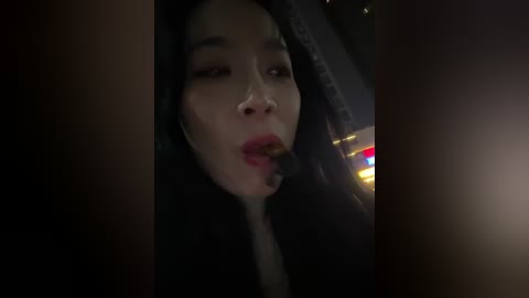 A dimly lit video captures an Asian woman with long black hair, wearing dark clothing, and a cigarette in her mouth, sitting in a car at night.