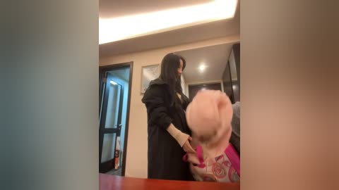 Media: A video of a woman in a black coat holding a child's hand in a brightly lit hallway, with a mirror reflecting their faces and a blurred doorway in the background.