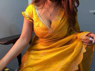 Video of a woman with medium brown skin, wearing a yellow saree with intricate embroidery and a gold necklace, sitting on a wooden chair against a plain gray wall.