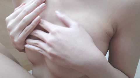 A close-up video shows a light-skinned person with small breasts, hands covering them. The background is blurred, featuring neutral tones, possibly a bathroom or bedroom setting.