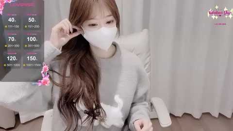 A video of an Asian woman with long brown hair, wearing a gray sweater and white mask, adjusting her hair in front of a white curtain backdrop.