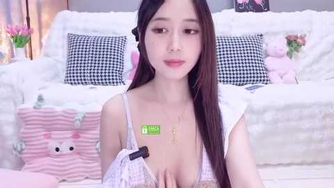 Video of an East Asian woman with long black hair, wearing a pink and white striped bra, sitting on a plush bed with white and black pillows.