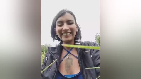 Video of a woman with a medium complexion, short black hair, and a broad smile, wearing a black jacket over a blue top, outdoors in a blurred, overcast landscape.