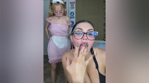 A video of a woman in a maid costume, standing in a room, with another woman, dressed casually, kneeling and eating a finger.