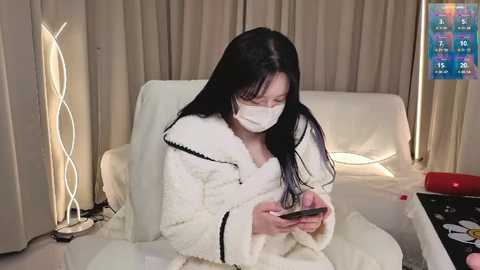 Video of an East Asian woman in a white robe with a black trim, wearing a white mask, playing a smartphone game in a modern living room with beige curtains, a white sofa, and a colorful digital clock.
