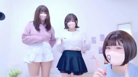 Video of three East Asian women in a brightly lit room. One wears a pink sweater, another a white sweater, and the third a blue top. All have short hair, with one wearing a mask.