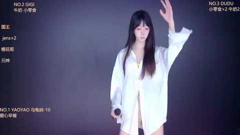 Video of an Asian woman with long, straight black hair, wearing a white lab coat and holding a microphone, against a dark background.