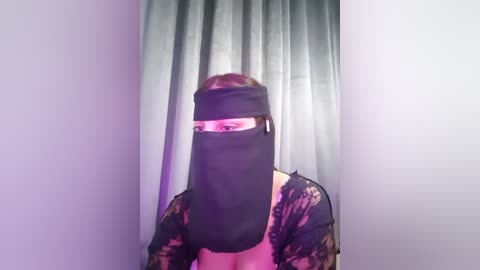 Video of a woman with light skin, wearing a black face veil and a sheer, lace top, standing in front of gray curtains.