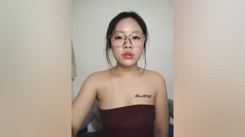 Video of an Asian woman with light skin, wearing glasses, and a strapless brown dress, with \"My Love\" tattoo on her shoulder, against a plain white wall.