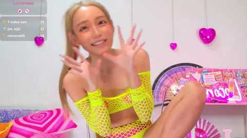 Video of a blonde, slim young woman with fair skin, wearing a neon yellow fishnet top, sitting in a colorful, heart-decorated room with pink walls and toys.