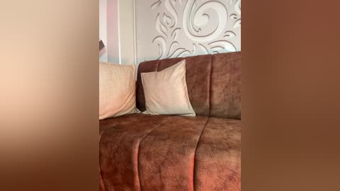 A video of a cozy, brown suede sectional sofa with two beige cushions against a textured, ornate, cream-colored wall. The background includes a hint of a white doorframe and soft lighting.