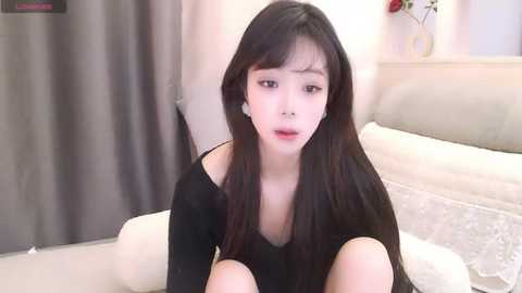 A video of a young East Asian woman with long black hair, fair skin, and red lipstick, wearing a black off-shoulder top, sitting on a beige couch in a softly lit, minimalist room.
