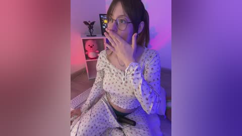 Video of a young woman with glasses, wearing white pajamas with a skull pattern, taking a selfie in a room with purple and pink lighting, a skull figurine on a shelf, and a plush toy.