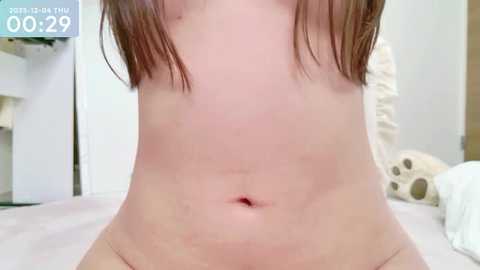 A close-up video of a light-skinned person with a flat stomach, featuring a small, red, circular mark on the abdomen, taken in a bedroom with white sheets and a beige pillow in the background.