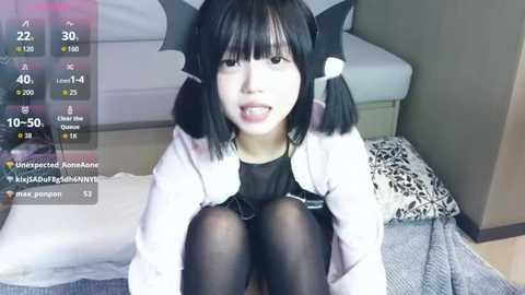 Video of a young Asian woman with straight black hair in bat wings, wearing a black dress and sheer black tights, sitting on a bed with white and patterned pillows.