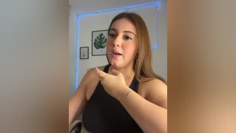 Video of a young, light-skinned woman with straight, light brown hair, wearing a black top. She's indoors, with a white wall, a plant, and blue LED lights in the background. She's smiling and making a peace sign with her hands.