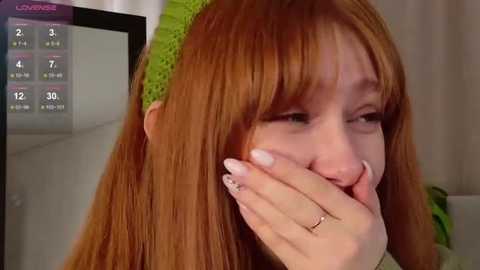 Video of a fair-skinned woman with long, straight, reddish-brown hair, wearing a lime green headband, covering her mouth with her hand, in a modern living room with a TV screen showing a weather forecast.