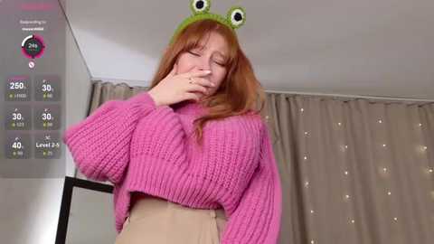Video of a red-haired woman with a green frog hat and pink knit sweater, standing in a dimly lit room with beige curtains.