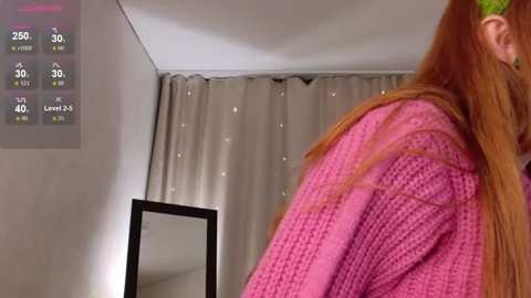 Video of a young woman with long red hair, wearing a pink sweater, in a modern, minimalist bedroom with a digital display and a mirror.