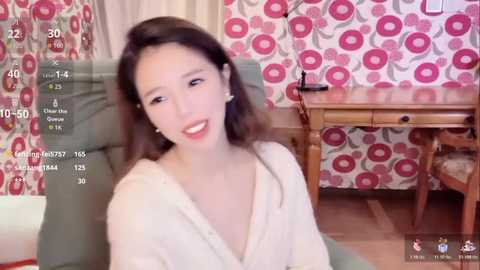 A video of an Asian woman with long black hair and light skin, wearing a white robe, sitting in a room with pink and white floral wallpaper and a wooden desk.