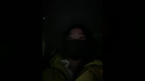 A dimly lit video of a person with dark skin, wearing a black mask and a yellow jacket, partially visible in the frame. The background is dark and blurry.