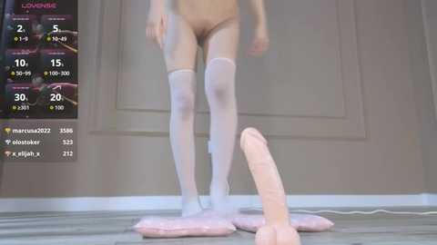 Video of a nude woman with fair skin wearing white thigh-high stockings, standing on a pink fur rug. Background shows a dimly lit room with a wooden floor and white walls.