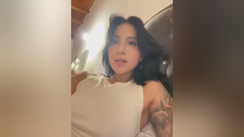 A video of a young Asian woman with medium-length black hair, wearing a white sleeveless top, sitting in a cozy indoor setting. She has a tattoo on her right arm. The background features wooden beams and a bed with patterned sheets.