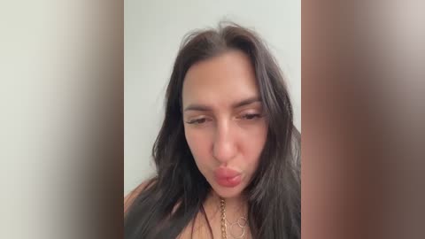 Video of a young woman with long dark hair, light brown skin, and full lips, puckering them, wearing a gold necklace, against a plain white background.