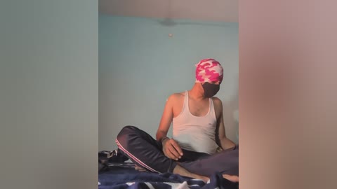 Video of a person with dark skin, wearing a pink and white floral headscarf, light tank top, and dark pants, sitting cross-legged on a bed with a blue and white patterned blanket, in a simple, sparsely decorated room with light blue walls.