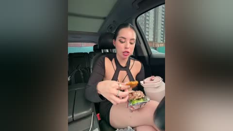 Video of a young woman with long dark hair, wearing a black halter top, eating a burger in the backseat of a car, blurred background.