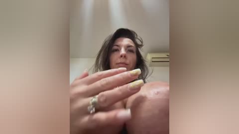 Media: A low-angle video of a middle-aged woman with long dark hair, light skin, and prominent breasts, wearing a gold ring. The background shows a beige wall with an air conditioner.
