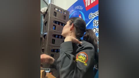 Video of a woman with dark hair tied back, wearing a black hoodie with a yellow logo, standing in front of a blue and white promotional poster, next to a tall, brown cardboard display.