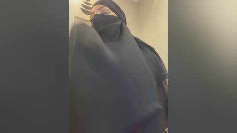 Video of a woman in a black burqa, partially obscured, with a blurred background featuring beige walls and a ceiling.